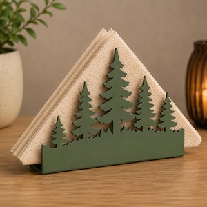May include: A green metal napkin holder with a forest silhouette design. The holder features a row of stylized trees and holds a stack of beige napkins. The holder is on a wooden surface.