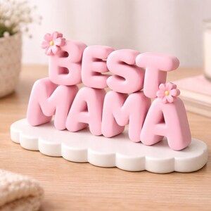 May include: A pink "BEST MAMA" decorative sign with flower accents, set on a white cloud-shaped base. The sign is displayed on a wooden surface, with a candle and flowers in the background.