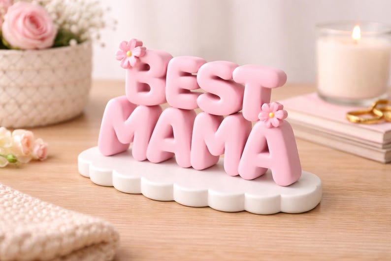 May include: A pink "BEST MAMA" decorative sign with flower accents, set on a white cloud-shaped base. The sign is displayed on a wooden surface, with a candle and flowers in the background.