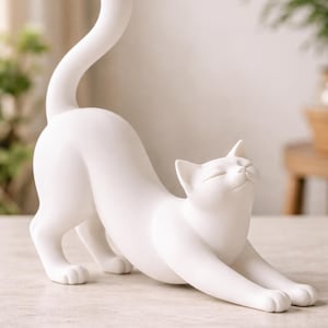 May include: A white ceramic cat ornament in a stretching pose. The cat has its eyes closed and its tail arched. The ornament is on a light-coloured surface, with a blurred background.