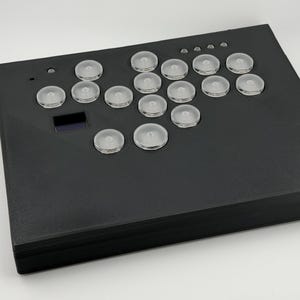 May include: A black arcade controller with clear, rounded buttons arranged in a grid. The rectangular controller has a small, rectangular screen. The buttons are evenly spaced across the top surface. The controller is designed for gaming.