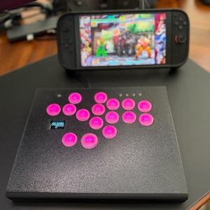 以下が含まれることがあります： ピンク色のボタンと小さなディスプレイを備えた、黒いアーケードスタイルのコントローラー。ゲーム画面を備えたハンドヘルドゲームデバイスが背景にあります。 コントローラーは黒い表面にあり、背景には木製の机があります。