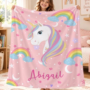 Op de afbeelding: Een zachte roze fleece deken met een centrale witte eenhoorn met een veelkleurige manen en hoorn. Het patroon bevat regenbogen, wolken, sterren, maansikkels en bloemen. De naam Abigail is onderaan in een cursief lettertype gedrukt.