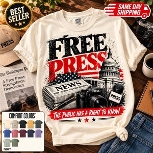 May include: A cream-colored short-sleeve t-shirt featuring a graphic print with the text FREE PRESS. The design includes a newspaper, a camera, a microphone labeled PRESS, and the US Capitol building, with the text THE PUBLIC HAS A RIGHT TO KNOW.
