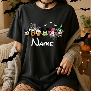 May include: A black short-sleeved t-shirt featuring a graphic print of six cartoon character heads in Halloween costumes, including a witch hat and spider webs. The word Name is printed in white below the characters. The shirt has a crew neckline.