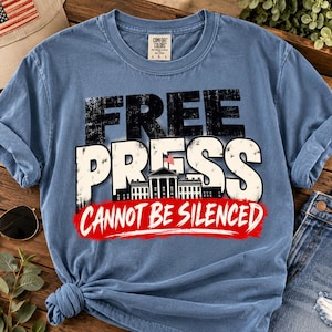Può includere: Una t-shirt blu a maniche corte con una stampa grafica che presenta il testo "FREE PRESS" in bianco e nero, un'illustrazione della Casa Bianca con una piccola bandiera americana e il testo rosso "CANNOT BE SILENCED" nella parte inferiore.