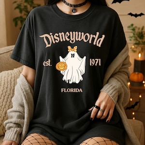 Könnte beinhalten: Ein schwarzes Kurzarm-T-Shirt mit dem Schriftzug Disneyworld est. 1971 FLORIDA in hellpfirsichfarbenen Buchstaben. Die Grafik in der Mitte zeigt ein weißes Gespenst mit einer orangefarbenen gepunkteten Schleife, das einen Kürbis hält.