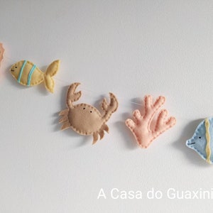 May include: A felt garland with sea creatures, including a seahorse, fish, crab, coral, and a blue angelfish. The garland is strung on a white string and has the text "A Casa do Guaxinim" at the bottom.