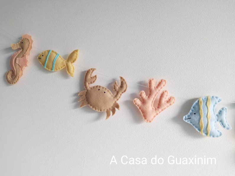 May include: A felt garland with sea creatures, including a seahorse, fish, crab, coral, and a blue angelfish. The garland is strung on a white string and has the text "A Casa do Guaxinim" at the bottom.