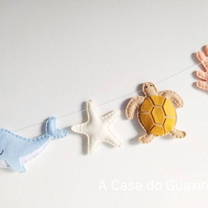 May include: A felt garland with a blue whale, a white starfish, a yellow turtle, and a pink coral. The garland is strung on a white string and has the text "A Casa do Guaxinim" at the bottom.