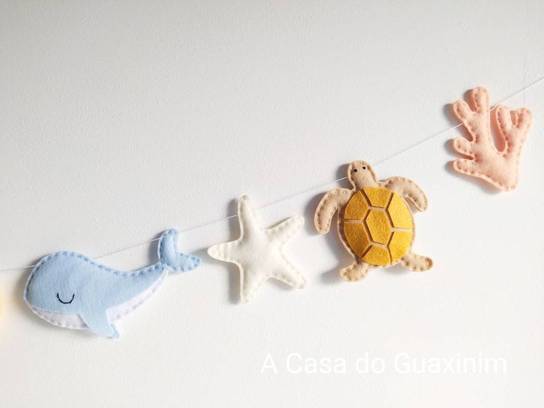 May include: A felt garland with a blue whale, a white starfish, a yellow turtle, and a pink coral. The garland is strung on a white string and has the text "A Casa do Guaxinim" at the bottom.