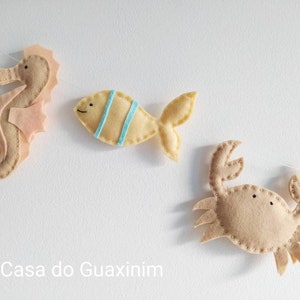 May include: A felt garland with a seahorse, fish, and crab. The seahorse is pink and brown, the fish is yellow with blue stripes, and the crab is brown.