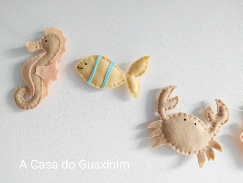 May include: A felt garland with a seahorse, fish, and crab. The seahorse is pink and brown, the fish is yellow with blue stripes, and the crab is brown.