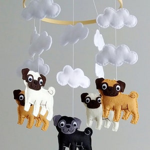Op de afbeelding: Een babymobiel met een houten ring en witte wolken van vilt. Er hangen vijf viltmoppen aan de ring, één zwarte, één bruine, één witte en twee bruine. De mobiel is gelabeld met "A Casa do Guaxinim".