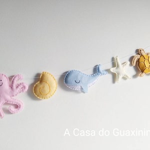 May include: A felt garland with a pink octopus, a yellow seashell, a blue whale, a white starfish, a yellow turtle, and a coral. The garland is strung on a white background. A Casa do Guaxinim is written below the garland.