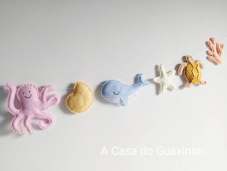 May include: A felt garland with a pink octopus, a yellow seashell, a blue whale, a white starfish, a yellow turtle, and a coral. The garland is strung on a white background. A Casa do Guaxinim is written below the garland.