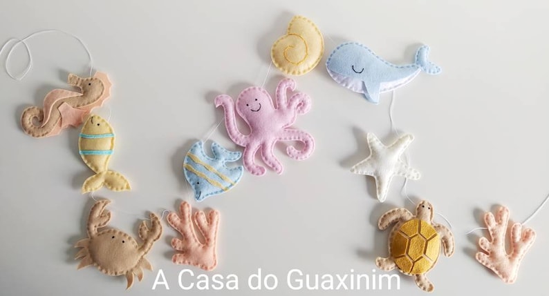 May include: A felt garland with a variety of sea creatures, including a seahorse, fish, octopus, starfish, turtle, and coral. The garland is strung on a white cord and is perfect for a nursery or child's room.
