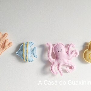 May include: A felt garland with a pink octopus, a blue fish, a coral, and a yellow seashell. The garland is strung on a white string. The text "A Casa do Guaxinim" is printed below the garland.