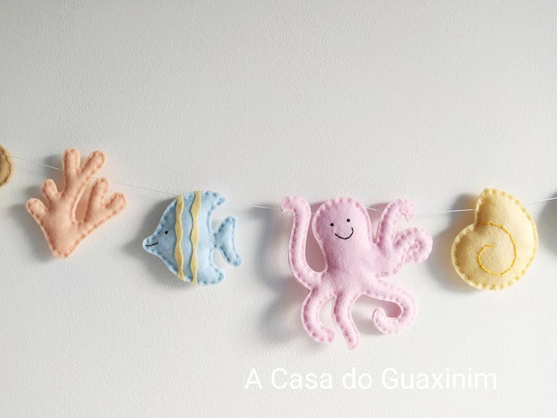 May include: A felt garland with a pink octopus, a blue fish, a coral, and a yellow seashell. The garland is strung on a white string. The text "A Casa do Guaxinim" is printed below the garland.