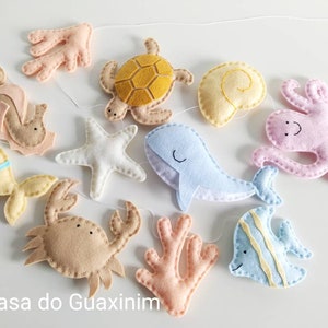May include: A felt ocean mobile with a variety of sea creatures, including a starfish, a seahorse, a crab, a turtle, a whale, an octopus, a seashell, and two fish. The mobile is made in pastel colors and is perfect for a nursery or child's room.