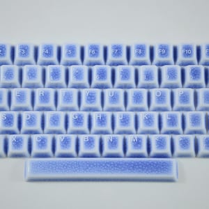 Pode incluir: Um conjunto de teclas de teclado de cerâmica azul claro e branco dispostas em um layout de teclado. As teclas têm um design salpicado e apresentam letras e números brancos. A barra de espaço também está incluída.