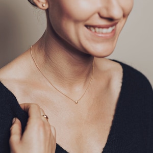 May include: A woman wearing a black sweater with a delicate gold chain necklace featuring a small, round pendant.