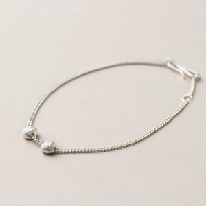 Op de afbeelding: Een delicate zilveren armband met twee getextureerde, bolvormige kralen. De armband heeft een fijne ketting en een eenvoudige sluiting. De sieraden worden gepresenteerd op een neutrale achtergrond.