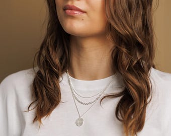 Op de afbeelding: Een vrouw draagt een wit T-shirt met een gelaagde zilveren ketting met een ronde hanger.