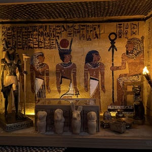 Puede incluir: Un diorama de caja de sombras de madera que representa una escena del antiguo Egipto. El interior presenta jeroglíficos pintados, figuras de faraones y dioses, y pequeñas estatuas. Dos pequeñas luces LED iluminan la escena, creando un brillo cálido.