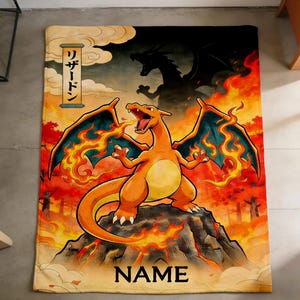 Może przedstawiać: Prostokątny koc z żywą ilustracją Charizarda, Pokemona typu ognistego, z płomieniami i sylwetką smoka w tle. Koc ma japoński tekst "リザードン" i słowo "NAME" na dole.