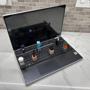Pode incluir: Um laptop preto com vários objetos em miniatura dispostos no teclado. Estes incluem uma xícara de café, uma garrafa, uma caneca azul, um cacto em um vaso e outros itens pequenos. O laptop está aberto, refletindo os objetos.