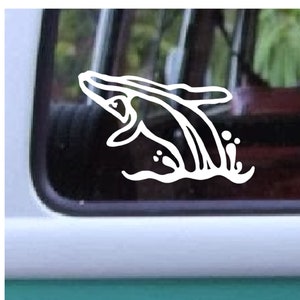 May include: White vinyl decal of a whale breaching the water. The whale is depicted with simple, clean lines, with water droplets and waves. The decal is applied to a window, with a blurred background of trees visible through the glass.