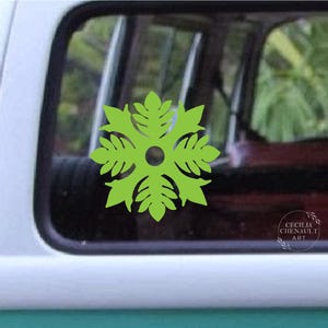 May include: A lime green leaf-shaped decal with a central hole, affixed to a vehicle window. The design features a symmetrical arrangement of leaf-like shapes radiating outwards. The decal is set against a backdrop of a vehicle interior and exterior.