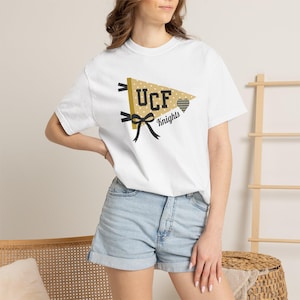Puede incluir: Camiseta blanca con un diseño de banderín dorado y negro. El banderín muestra el texto "UCF Knights" y un corazón. El banderín tiene un patrón de lunares y cintas negras en el lado izquierdo.