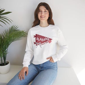 May include: White sweatshirt with a maroon pennant design. The pennant reads "ALABAMA" and "Crimson Tide". The design includes a polka dot pattern and a bow. The sweatshirt is long-sleeved.