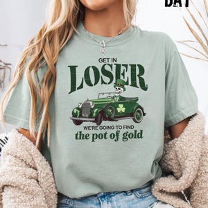 May include: Sage green t-shirt with the text "GET IN LOSER" above a green vintage car graphic. The car features a skeleton driver and a shamrock. The phrase "WE'RE GOING TO FIND the pot of gold" is below the car.
