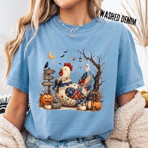 May include: A light blue t-shirt featuring a graphic of a patchwork chicken surrounded by pumpkins, a wooden signpost labeled BOO, TREATS, and PUMPKINS, a bare tree, a crescent moon, and a small ghost. The shirt has a crew neckline and short sleeves.