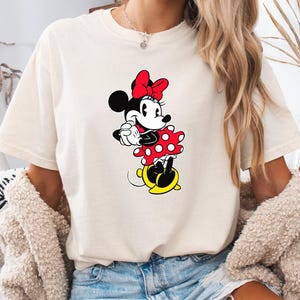 May include: Ivory-colored t-shirt featuring a Minnie Mouse graphic. The cartoon character is depicted in a red polka-dot dress, a red bow, and yellow shoes. The word "IVORY" is printed in black at the top left corner of the shirt.