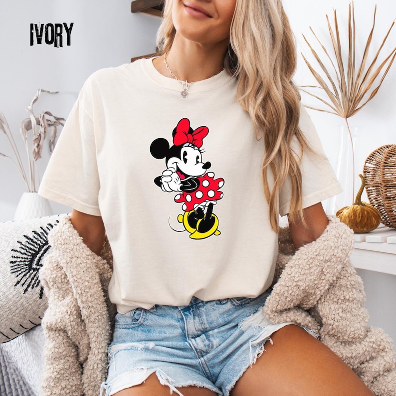 May include: Ivory-colored t-shirt featuring a Minnie Mouse graphic. The cartoon character is depicted in a red polka-dot dress, a red bow, and yellow shoes. The word "IVORY" is printed in black at the top left corner of the shirt.