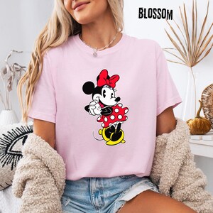May include: A light pink t-shirt featuring a Minnie Mouse graphic. The cartoon character is depicted in a red polka-dot dress and bow, with yellow shoes. The word "BLOSSOM" is printed in black at the top right.