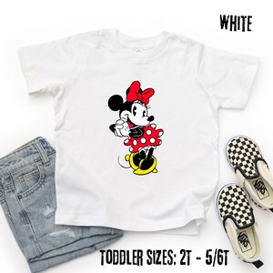 May include: White toddler t-shirt featuring a Minnie Mouse graphic. Minnie is depicted in her classic outfit: a red polka-dot dress, a red bow, and yellow shoes. The text "WHITE" is at the top right, and "TODDLER SIZES: 2T - 5/6T" is at the bottom.