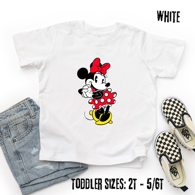 May include: White toddler t-shirt featuring a Minnie Mouse graphic. Minnie is depicted in her classic outfit: a red polka-dot dress, a red bow, and yellow shoes. The text "WHITE" is at the top right, and "TODDLER SIZES: 2T - 5/6T" is at the bottom.