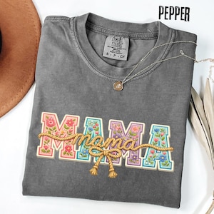 Może przedstawiać: Szara koszulka Comfort Colors z napisem "MAMA" w kolorowych kwiatowych literach, połączonych wzorem liny. Na koszulce znajduje się srebrny naszyjnik z małą zawieszką. U góry znajduje się napis "Pepper".