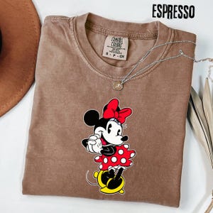 May include: Espresso-colored t-shirt featuring a Minnie Mouse graphic. The graphic shows Minnie in a red bow, red and white polka dot dress, and yellow shoes. The shirt has a Comfort Colors tag and a silver necklace.