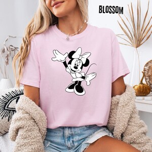 Peut inclure: T-shirt rose clair avec un dessin noir et blanc de Minnie Mouse, les bras lev&eacute;s. Le mot "BLOSSOM" est imprim&eacute; en noir en haut. Le t-shirt est fait d'une mati&egrave;re douce.
