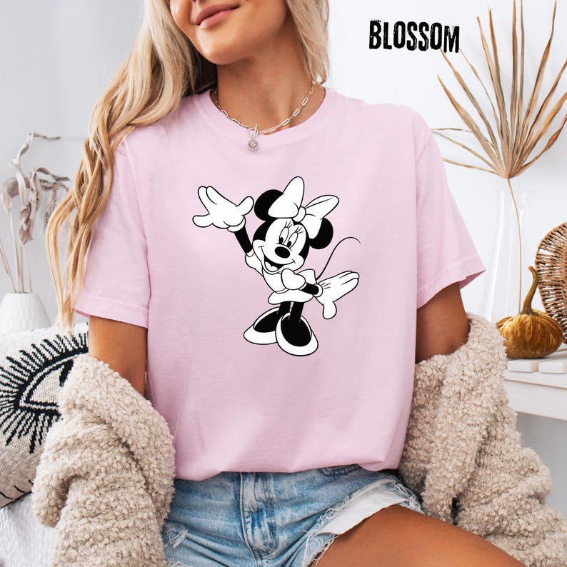 Peut inclure: T-shirt rose clair avec un dessin noir et blanc de Minnie Mouse, les bras lev&eacute;s. Le mot "BLOSSOM" est imprim&eacute; en noir en haut. Le t-shirt est fait d'une mati&egrave;re douce.