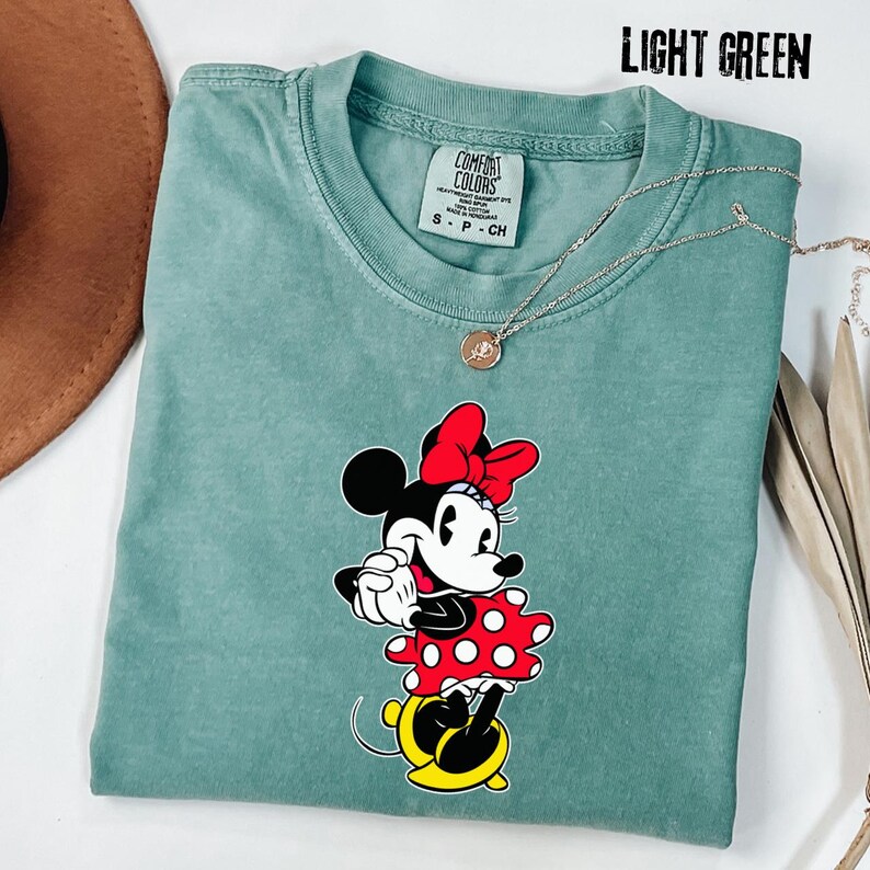 May include: Light green Comfort Colors t-shirt featuring a Minnie Mouse graphic. The graphic shows Minnie Mouse in a red bow, red and white polka dot dress, black ears, and yellow shoes. A silver necklace is draped over the shirt.