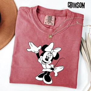 Peut inclure: T-shirt de couleur cramoisie avec un motif Minnie Mouse en noir et blanc. Le t-shirt a un col rond et des manches courtes. Un collier argent&eacute; avec un pendentif est drap&eacute; sur le t-shirt. Le t-shirt est &eacute;tiquet&eacute; "Comfort Colors" et taille S-P-CH.