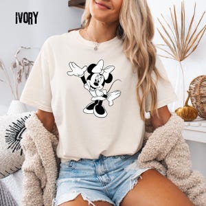 Peut inclure: T-shirt de couleur ivoire avec un dessin en noir et blanc de Minnie Mouse. Le personnage de dessin anim&eacute; est repr&eacute;sent&eacute; avec son n&oelig;ud et ses gants, dans une pose enjou&eacute;e. Le mot "IVORY" est imprim&eacute; en haut &agrave; gauche du t-shirt.