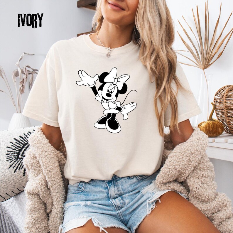 Peut inclure: T-shirt de couleur ivoire avec un dessin en noir et blanc de Minnie Mouse. Le personnage de dessin anim&eacute; est repr&eacute;sent&eacute; avec son n&oelig;ud et ses gants, dans une pose enjou&eacute;e. Le mot "IVORY" est imprim&eacute; en haut &agrave; gauche du t-shirt.